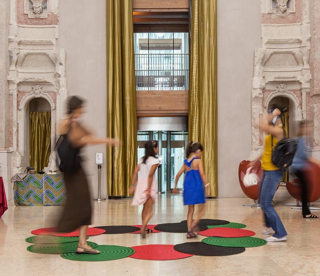 Duas mulheres e duas crianças andam à volta sob tapetes de várias cores (vermelho, verde e preto)}