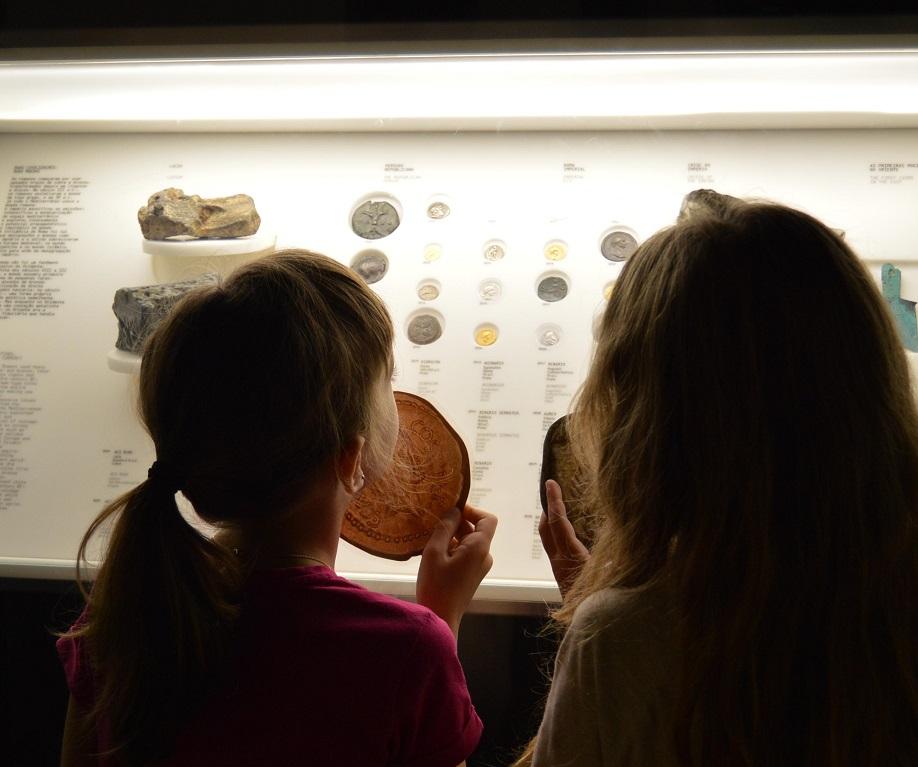 Duas crianças de costas espreitam para dentro de vitrine com moedas romanas